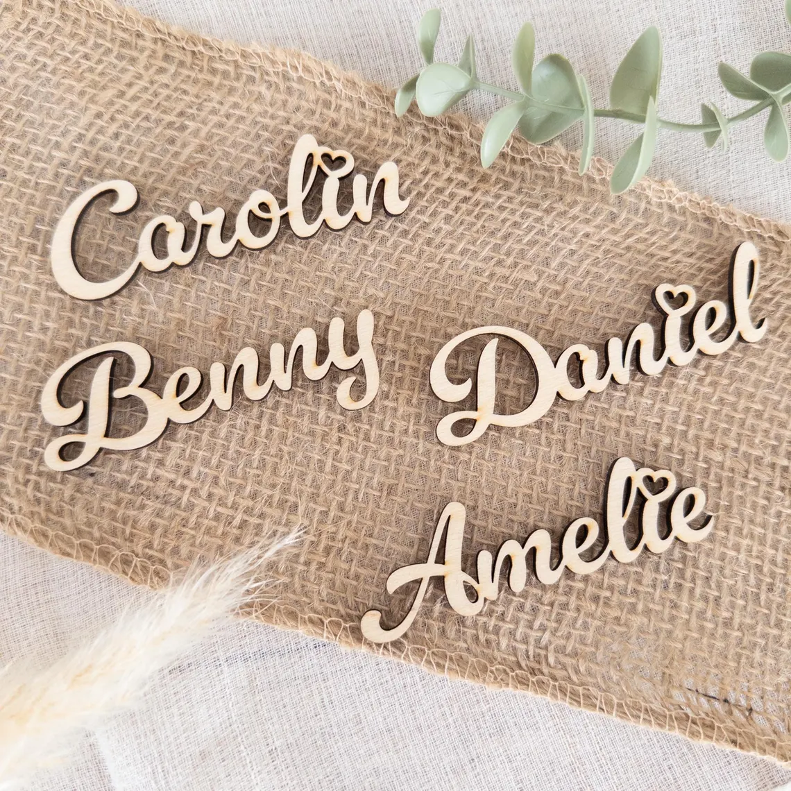 Vier filigrane, lasergravierte Namensschilder aus Holz mit den Namen Carolin, Benny, Daniel und Amelie liegen auf einem Stück Jutestoff und dienen als personalisierte Deko bzw. Platzkarten.