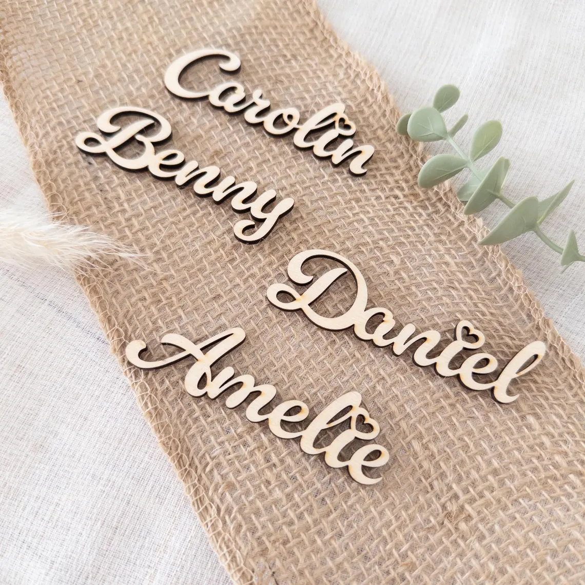 Vier filigrane, lasergravierte Namensschilder aus Holz mit den Namen Carolin, Benny, Daniel und Amelie liegen auf einem Stück Jutestoff und dienen als personalisierte Deko bzw. Platzkarten.