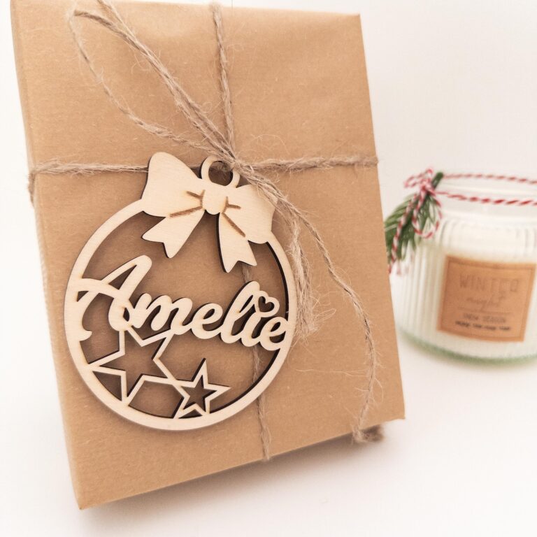 Nahaufnahme eines in Packpapier verpackten Geschenks, mit Juteschnur umwickelt und einem personalisierten, aus Holz gelaserten Weihnachtsanhänger mit dem Namen „Amelie“ und Sternmotiven; im Hintergrund steht eine Duftkerze.