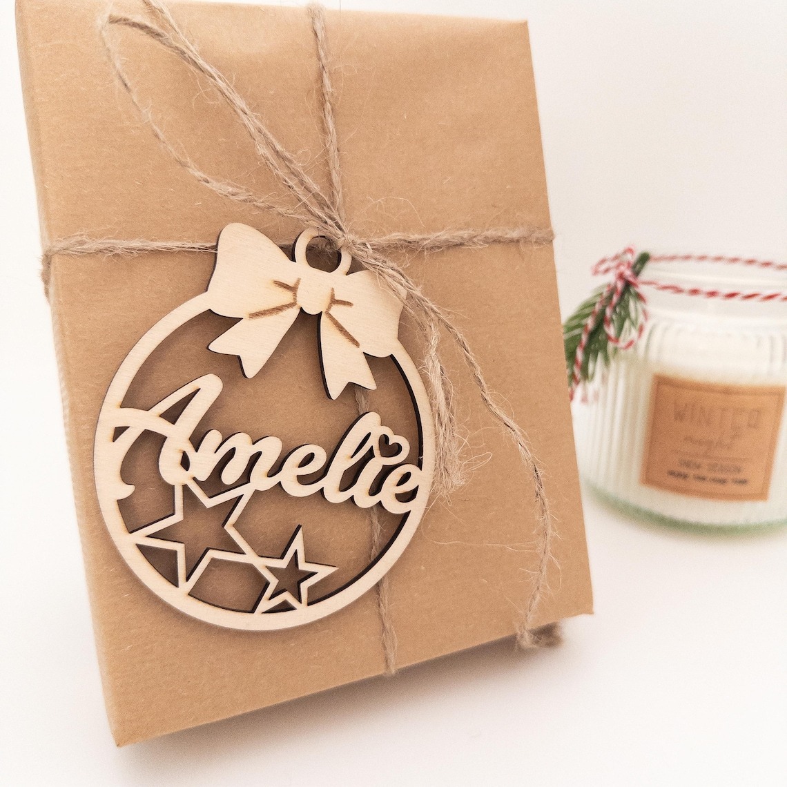 Nahaufnahme eines in Packpapier verpackten Geschenks, mit Juteschnur umwickelt und einem personalisierten, aus Holz gelaserten Weihnachtsanhänger mit dem Namen „Amelie“ und Sternmotiven; im Hintergrund steht eine Duftkerze.