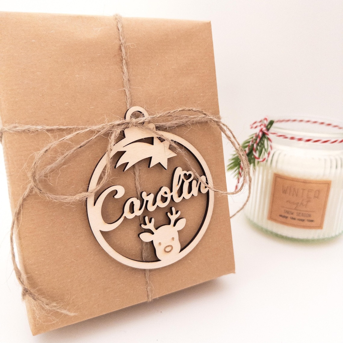 In Packpapier verpacktes Geschenk, mit Juteschnur verschnürt und einem personalisierten Holz-Weihnachtsanhänger mit dem Namen „Carolin“, Sternschnuppe und Rentiermotiv; im Hintergrund steht eine Winterduftkerze.