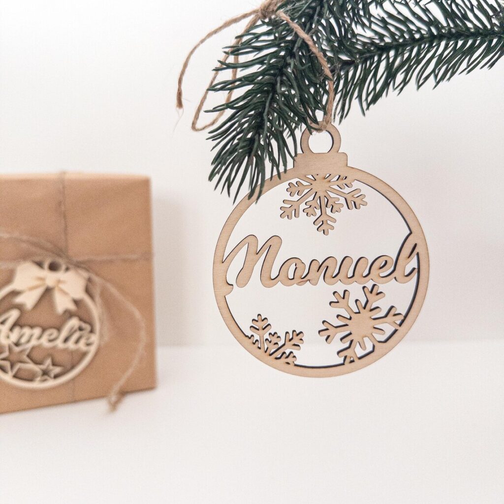 Personalisierter Weihnachtskugel aus gelasertem Holz mit dem Namen „Manuel“ und Schneeflockenmotiven hängt an einem Tannenzweig; im Hintergrund liegt ein in Packpapier verpacktes Geschenk mit weiterem Namensanhänger.