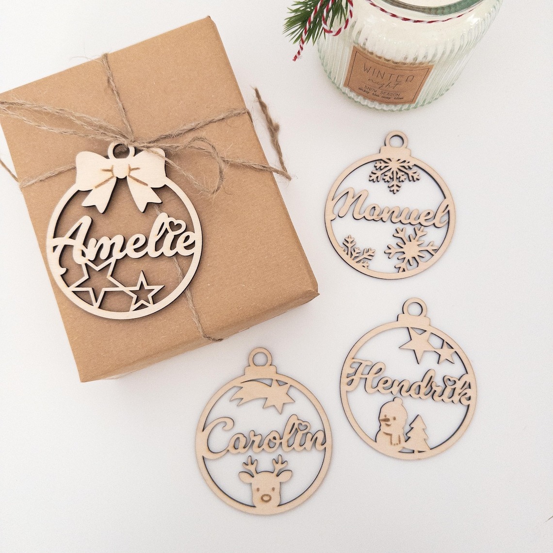 Vier personalisierte Weihnachtskugeln aus gelasertem Holz mit den Namen Amelie, Manuel, Carolin und Hendrik liegen neben einem schlicht verpackten Geschenk und einer Duftkerze auf einem weißen Untergrund.