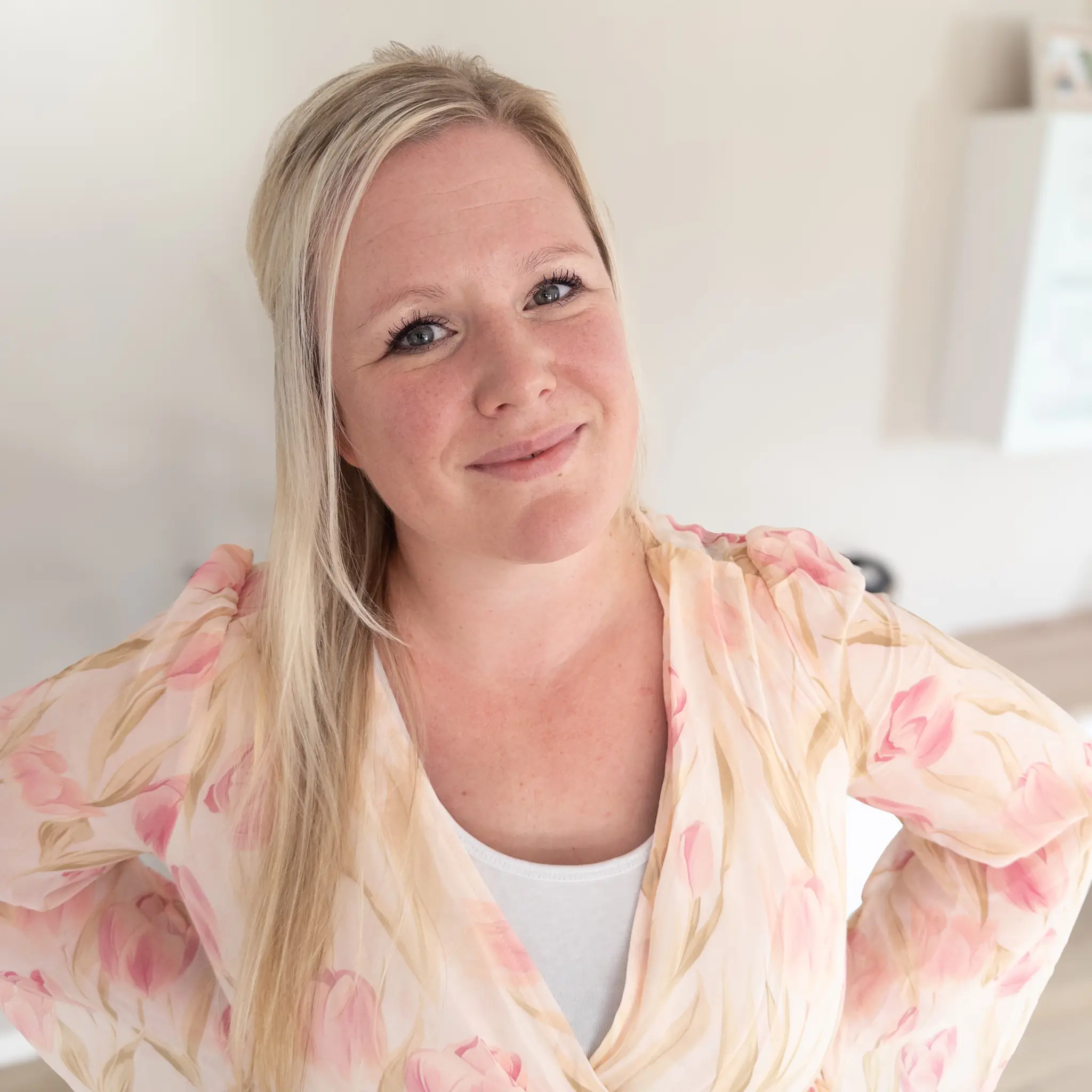 orträt einer lächelnden Frau mit blonden, halblangen Haaren in einem hellen, blumig gemusterten Kleid, die mit verschränkten Armen in einem freundlich wirkenden, hellen Raum steht.