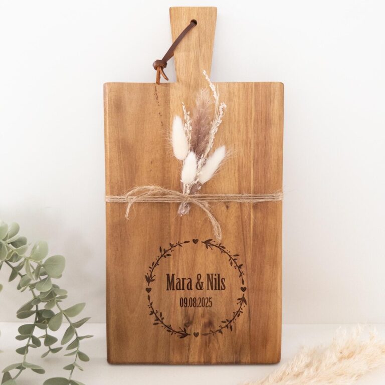 Personalisierte Schneidebrett mit eingraviertem Kranz, den Namen „Mara & Nils“ und dem Datum „09.08.2025“, mit Juteschnur und Trockenblumen dekoriert, daneben dezente Eukalyptus- und Pampasgräser.