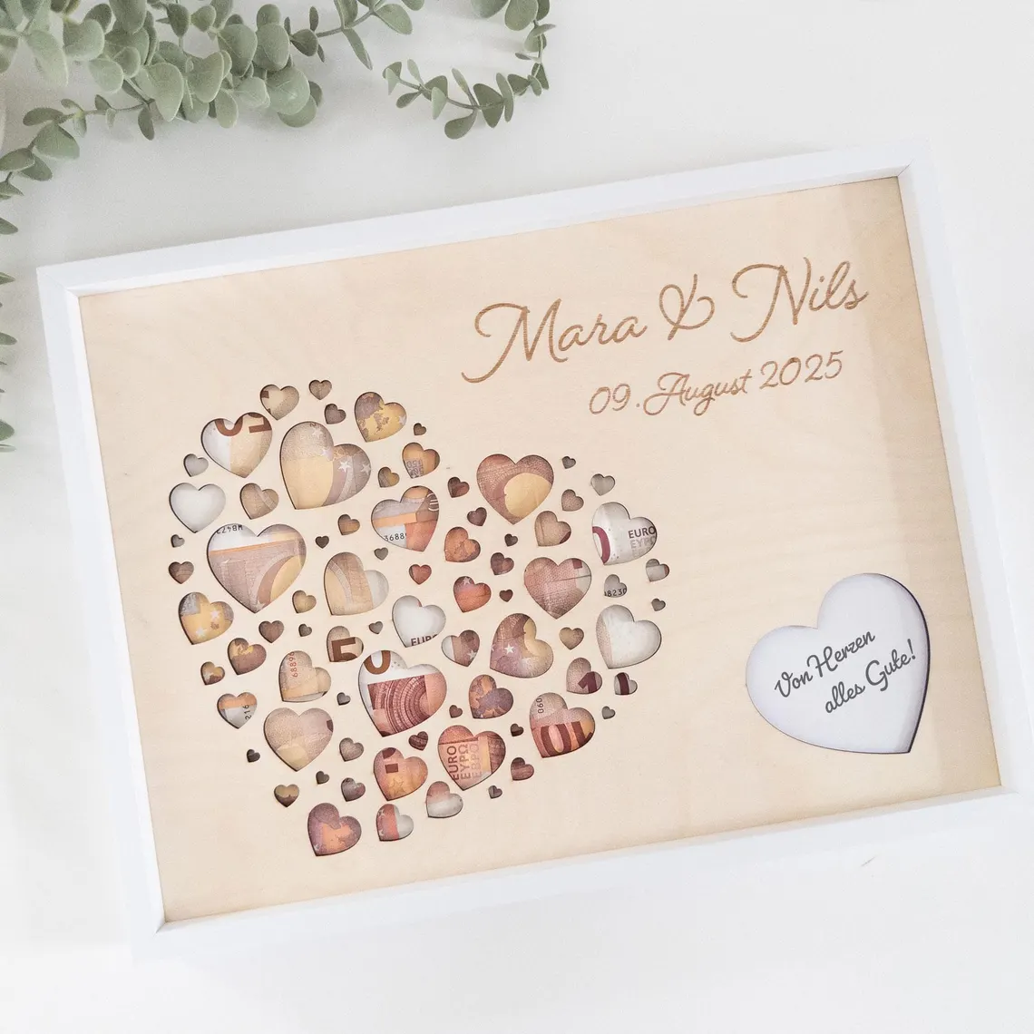 Ein Bilderrahmenmit einem herzförmigen Design, gefüllt mit Herz-Ausschnitten, einige davon mit Geld. Das weiß gerahmte Kunstwerk trägt die Namen "Mara & Nils" und das Datum "09. August 2025". Der Text "Von Herzen alles Gute!" ist ebenfalls sichtbar.