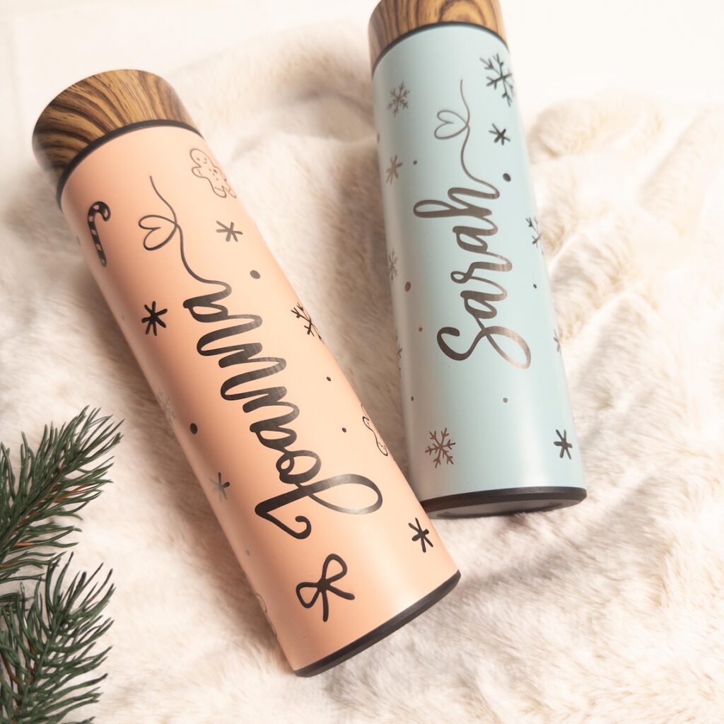Personalisierte Thermosflaschen aus Edelstahl in Pastellfarben mit Holzdeckel, bedruckt mit den Namen „Joanna“ und „Sarah“ sowie winterlichen Motiven wie Schneeflocken, Herzchen und Zuckerstange, dekorativ auf weißem Fell neben einem Tannenzweig arrangiert.