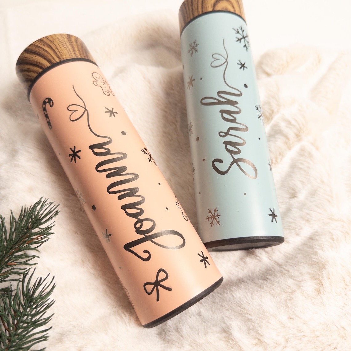 Personalisierte Thermosflaschen aus Edelstahl in Pastellfarben mit Holzdeckel, bedruckt mit den Namen „Joanna“ und „Sarah“ sowie winterlichen Motiven wie Schneeflocken, Herzchen und Zuckerstange, dekorativ auf weißem Fell neben einem Tannenzweig arrangiert.