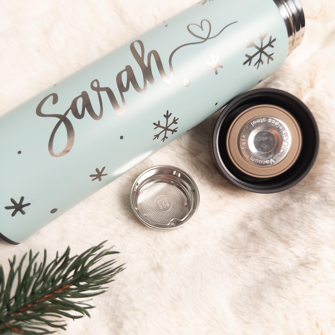 Personalisierte Türkis Thermosflasche aus Edelstahl mit Namen „Sarah“ und Schneeflocken-Motiven, liegend auf weißem Fell, daneben der abgenommene Deckel und ein herausnehmbarer Teesieb-Einsatz aus Edelstahl, dekoriert mit einem Tannenzweig im Vordergrund.
