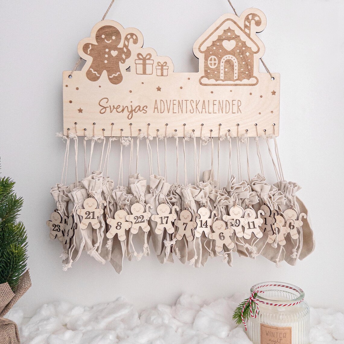 Holz-Adventskalender mit Lebkuchen-Motiven und Schrift „Svenjas Adventskalender“; darunter 24 beige Stoffbeutel mit nummerierten Anhängern; winterliche Deko und Glas im Vordergrund.