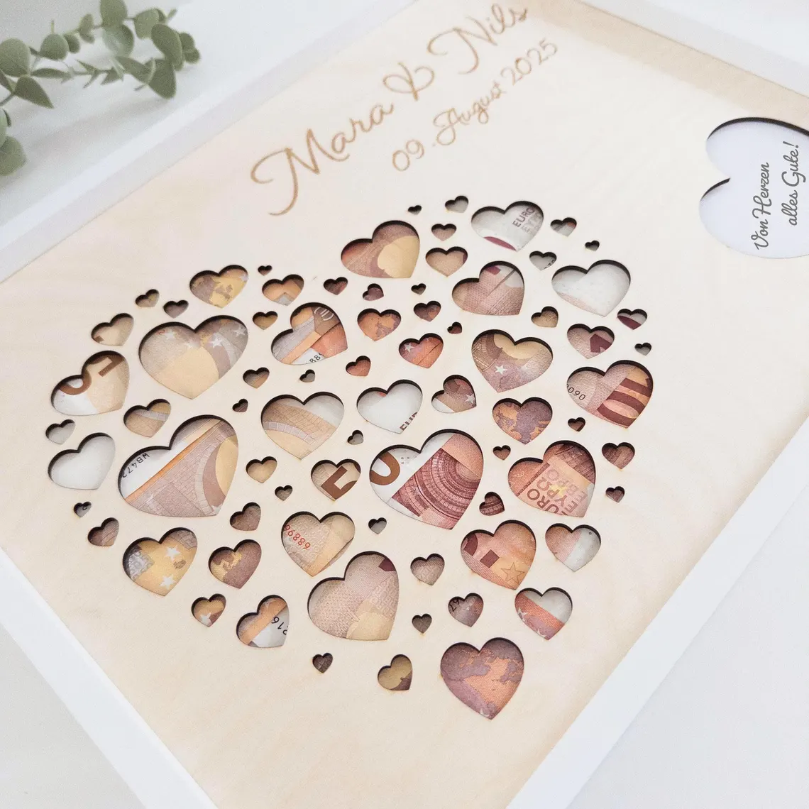 Ein Bilderrahmen mit einem herzförmigen Design, gefüllt mit herzförmigen Ausschnitten. Das Herz ist mit Währung gefüllt. Das Kunstwerk trägt die Namen "Mara & Nils" und das Datum "09 August 2025". Ein kleineres Herz mit Text ist ebenfalls vorhanden.