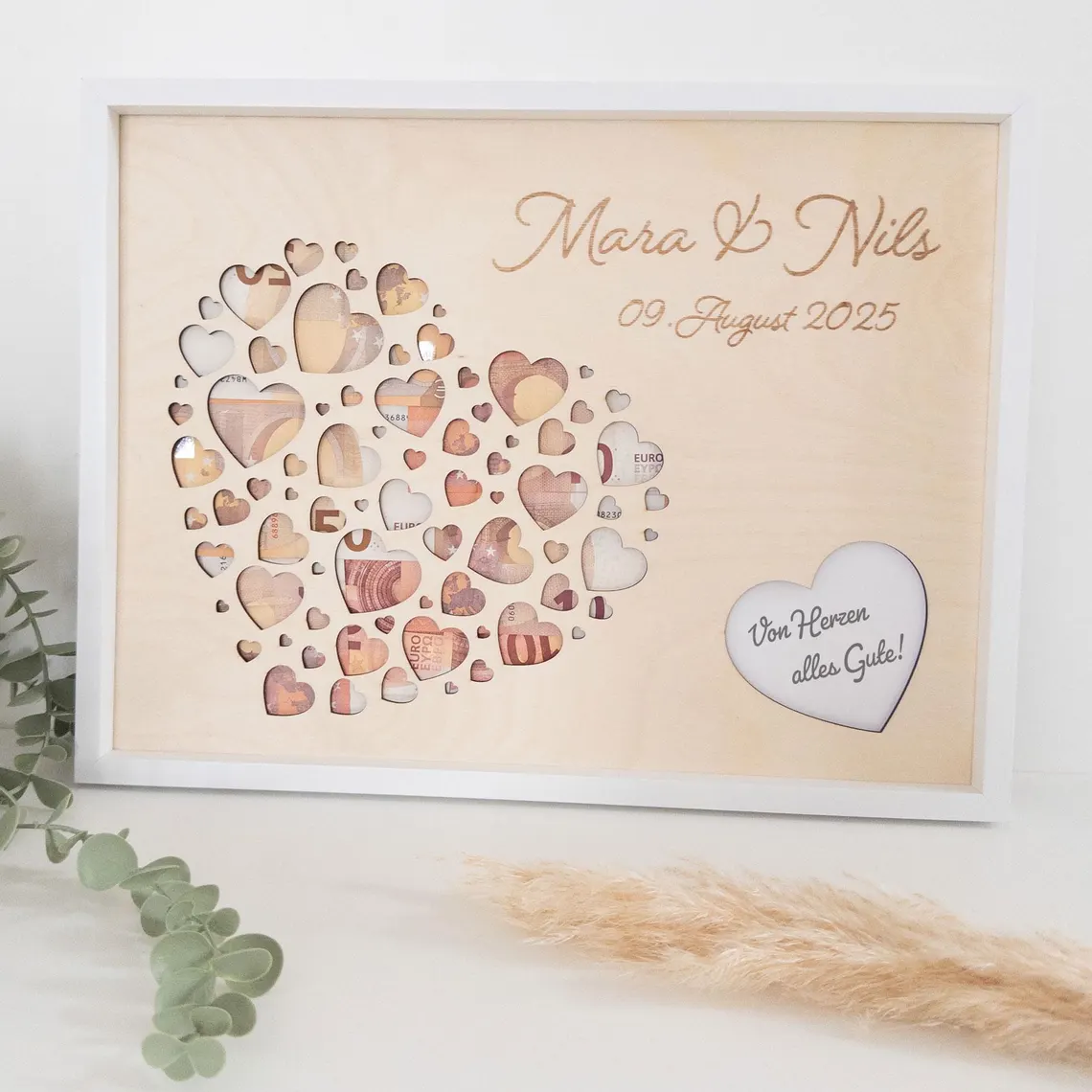 Ein Bilderrahmen mit einem herzförmigen Design, gefüllt mit herzförmigen Ausschnitten. Das Herz ist mit Währung gefüllt. Das Kunstwerk trägt die Namen "Mara & Nils" und das Datum "09 August 2025". Ein kleineres Herz mit Text ist ebenfalls vorhanden.