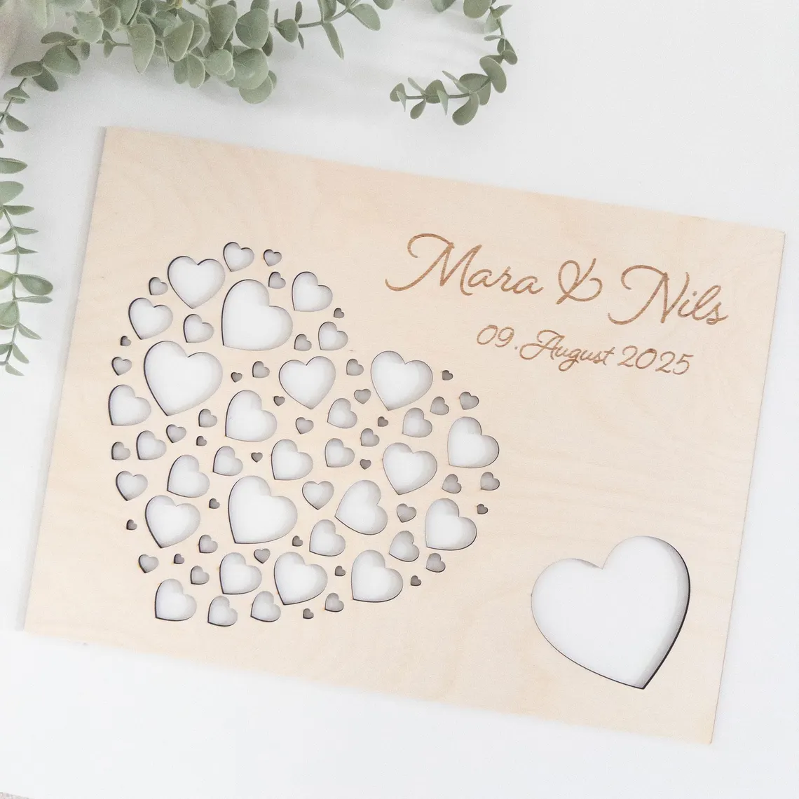 Helle Holzplatte mit einem großen herzförmigen Ausschnitt, der mit kleineren Herz-Ausschnitten gefüllt ist. Die Namen "Mara & Nils" und das Datum "09. August 2025" sind in das Holz graviert. Ein einzelner Herz-Ausschnitt befindet sich in der unteren rechten Ecke.