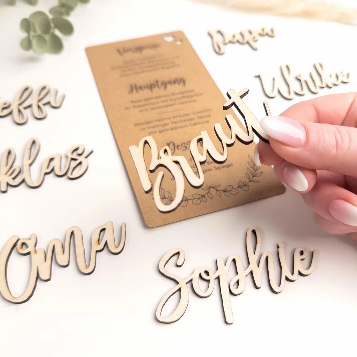 Holz-Namensschilder und eine Menükarte. Das Wort "Braut" wird von einer Hand gehalten. Weitere Namen sind "Sophie", "Oma" und "Klaus". Die Menükarte hat deutschen Text. Die Etiketten sind hellbraun.