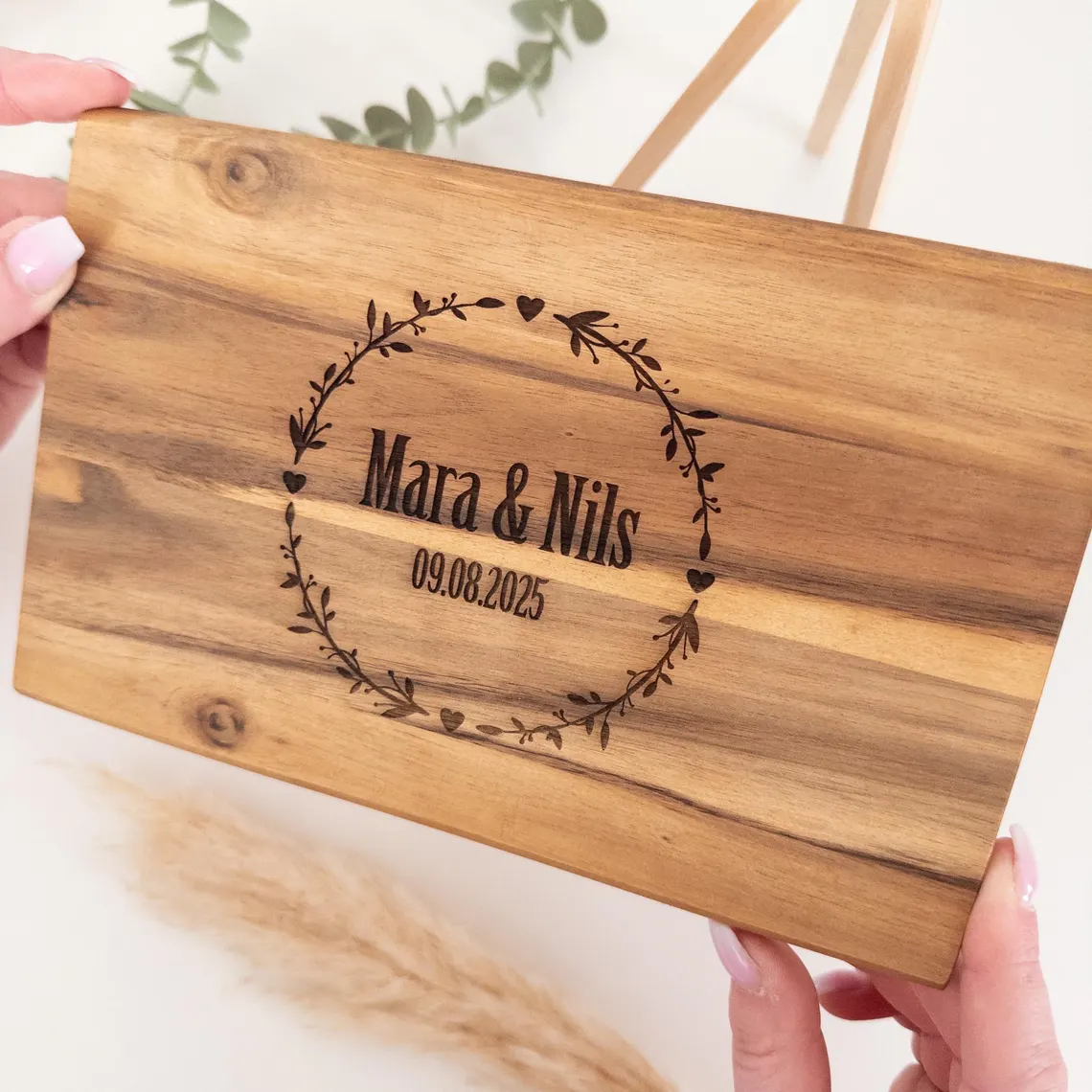 Nahaufnahme: personalisiertes Schneidebrett als Hochzeitsgeschenk, von zwei Händen gehalten; Gravur ‚Mara & Nils‘ mit Datum im Kranz, helle Deko mit Eukalyptus & Pampas.