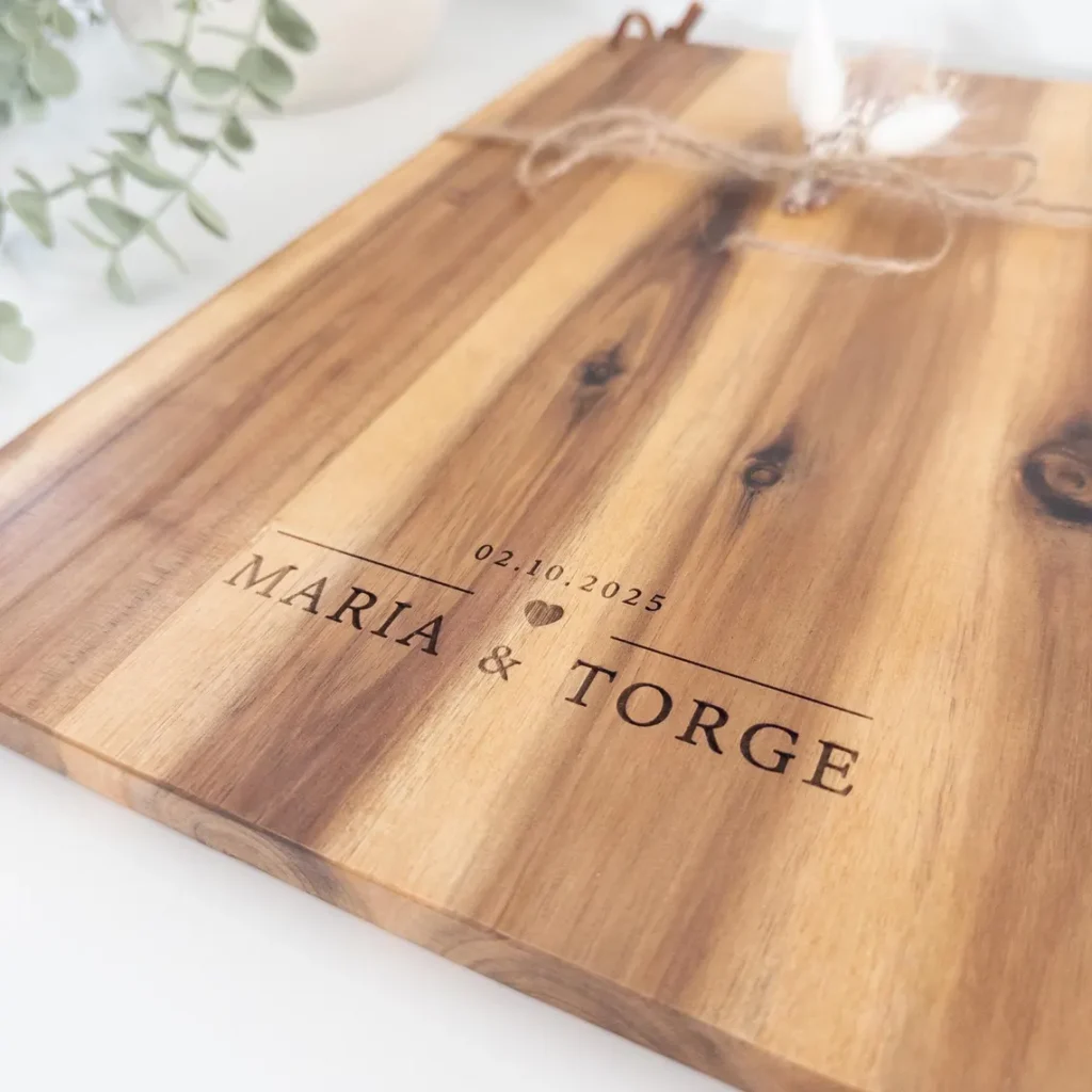 Nahaufnahme eines personalisierten Schneidebretts aus Akazienholz mit Lasergravur „Maria & Torge“ und Datum „02.10.2025“.